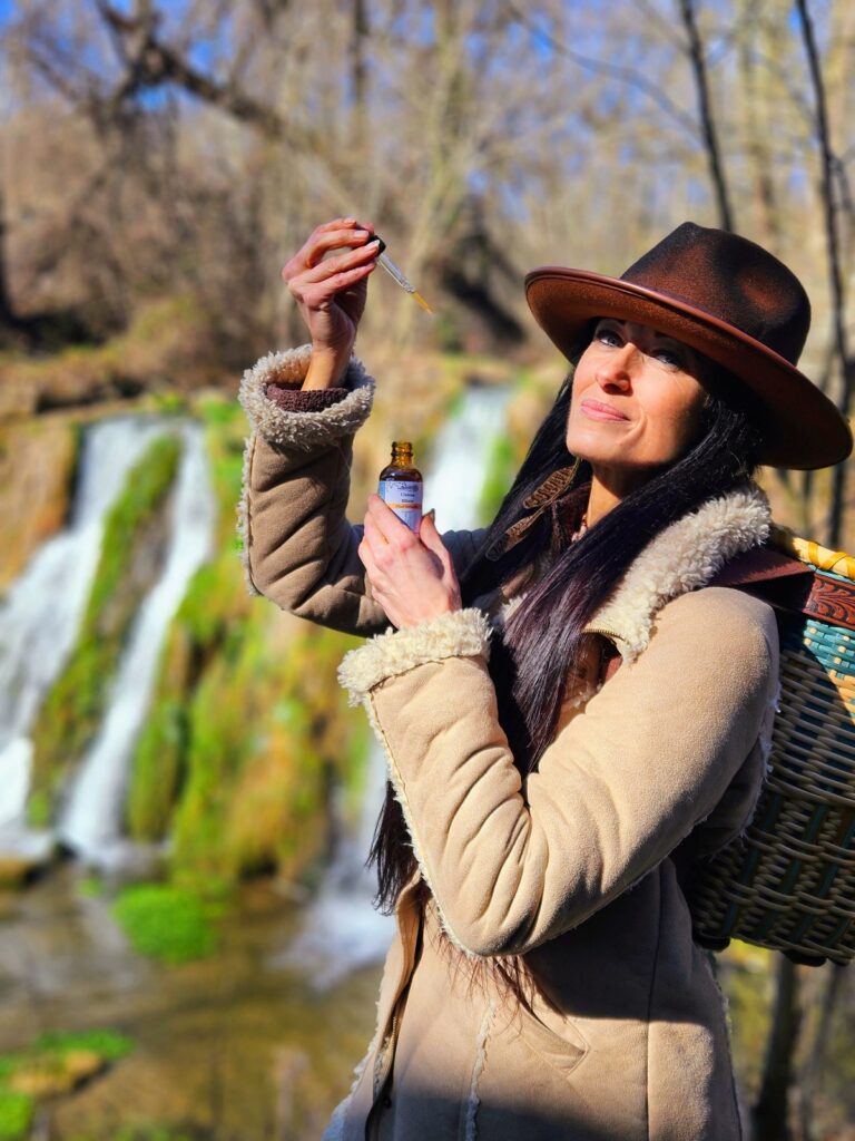 A herbalist woman in a hat holding a bottle of essential oil in front of a waterfall.